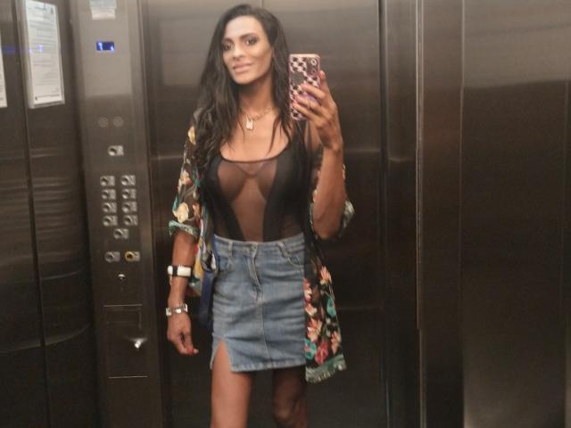 DiannaBanana with medium tits, wearing a sheer black top, floral kimono, and blue denim skirt, taking a mirror selfie in an elevator. Her phone has a pink, checkered case.