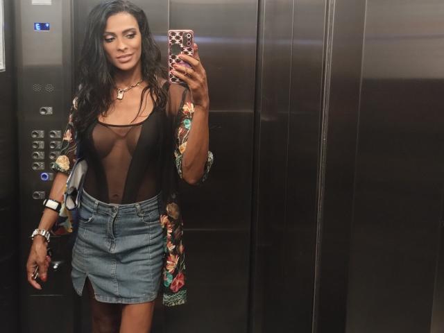 DiannaBanana with long black hair, wearing sheer black top showing her tits, colorful robe, blue denim skirt, and gold jewelry, taking a mirror selfie in an elevator.