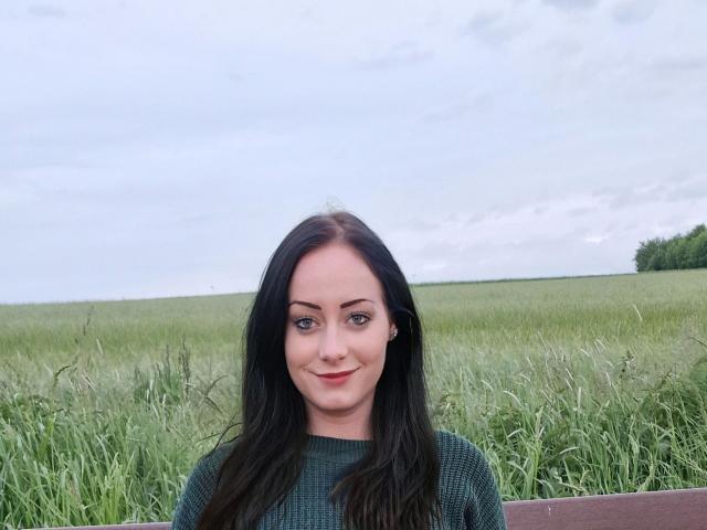JasminWhite-hot with long black hair, green eyes, and red lipstick, smiling in a green field, wearing a green knit sweater, standing in front of a wooden fence.