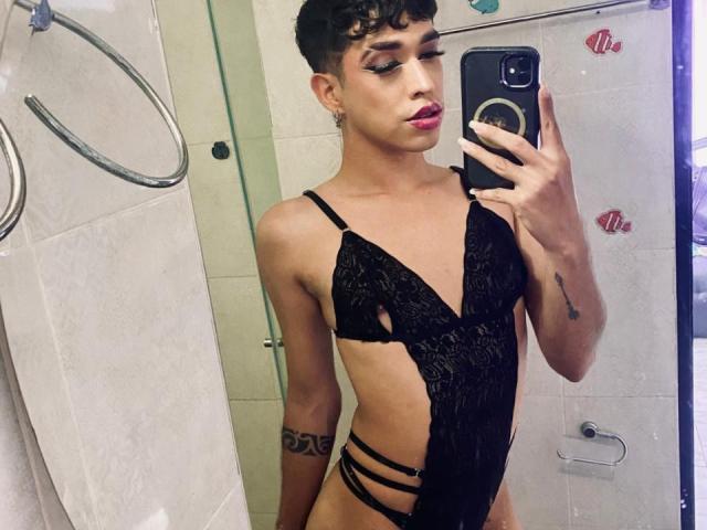 BriggitteLove met kort krullend haar, gekleed in zwarte kant lingerie, waarbij haar kleine borsten en smalle bandjes zichtbaar zijn, maakt een selfie in de spiegel met haar telefoon..