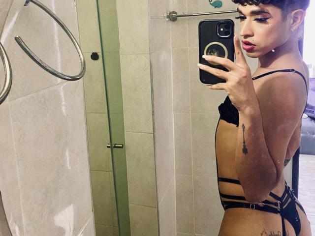 BriggitteLove with short brown hair, pouty pink lips, and black strappy lingerie, holding a phone in a bathroom mirror selfie, showing a tattoo on her right arm.