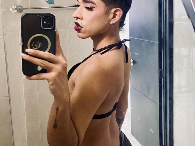 BriggitteLove with short dark hair, red lipstick, and black string bikini, holding phone, showing side profile, small tits, tattoo on arm, mirror selfie in bathroom.