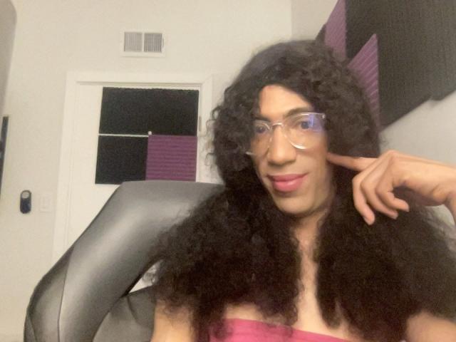 TehaniBeny with curly black hair, glasses, pink lipstick, and a pink strapless top, pointing to her cheek, sitting in a black chair.