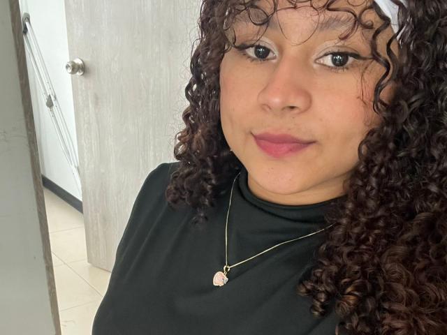SofiaPassionne with curly brown hair, pink lipstick, black turtleneck, gold necklace with pink gem, white headband, smiling, pale skin, medium breasts, brown eyes, indoors.