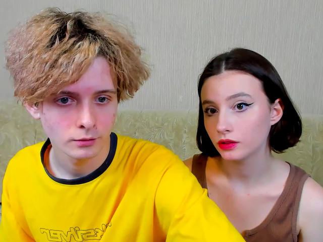 Michelle ha i capelli biondi e ricci, una camicia gialla e un'espressione seria. James ha i capelli castano scuro, rossetto rosso e una canotta marrone, con un'espressione neutra..