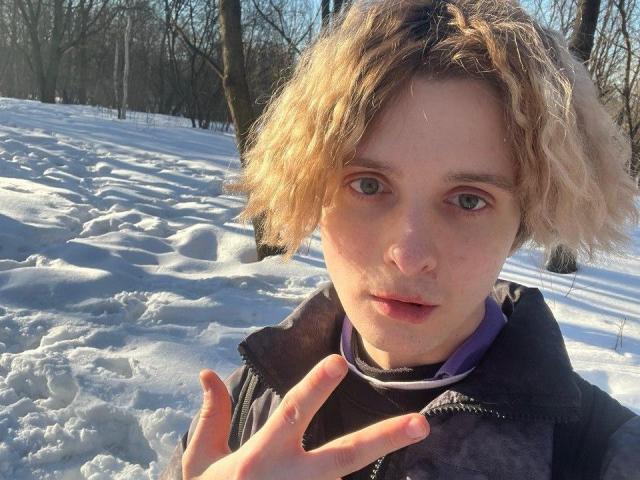 Michelleandjames with curly blonde hair, pale skin, and blue eyes, wearing a dark jacket, making a hand gesture in a snowy forest. Sunlight highlights her face.