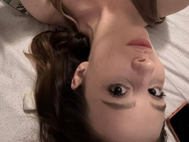 Michelleandjames's face, upside down, with brown eyes, eyebrows, and brown wavy hair, wearing a brown top, on a white textured surface, looking at the camera.