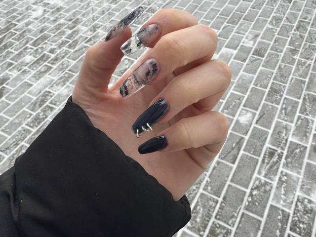Michelleandjames' hand with long, black and silver acrylic nails featuring abstract patterns, holding a clear, rectangular nail file, wearing a black sleeve, on a cobblestone surface.