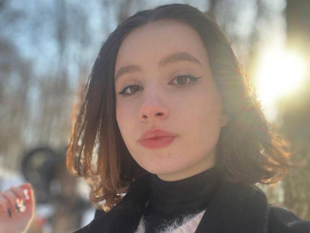 Michelleandjames has pale skin, dark brown shoulder-length hair, and black eyeliner with bold red lipstick. She's wearing a black turtleneck with a white furry collar. Sunlight flares.