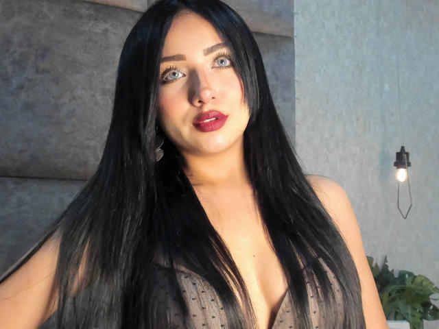 LillyTaylor has long black hair, blue eyes, and red lipstick. She's wearing a sheer, dark gray top that reveals her boobs. She's looking up with a slight smile.