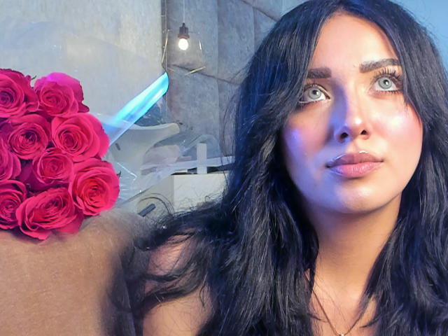 LillyTaylor with long black hair, blue eyes, and pinkish skin, holding a bouquet of red roses, her pouty lips slightly parted, looking up with a serious expression.