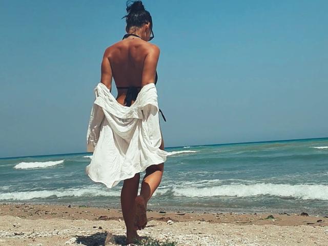 A photograph of a beach. HotBisous stands with her back to the camera, wearing a black bikini and a loose white shirt. Her dark hair is in a messy bun. She's walking on a sandy shore with small rocks and pebbles. The ocean is in the background with gentle waves and a clear blue sky. Her tan skin contrasts with the white shirt. The image focuses on her from behind, showing her bare back and bikini straps.