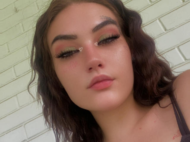 BarbyRH has wavy brown hair, green eyeshadow, heart-shaped eyeliner, long eyelashes, pink lips, and a slightly flushed face. She's looking down with a neutral expression.