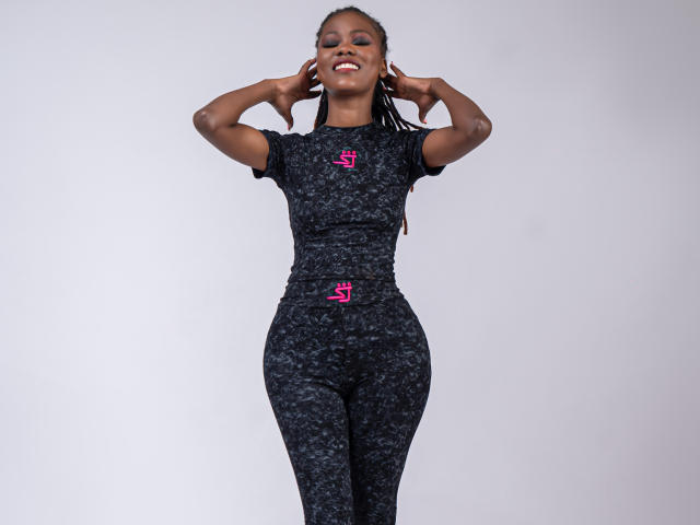 HotyBarbarha, dark-skinned, standing, smiling, hands behind head, wearing black, short-sleeve, textured, tight, athletic pants, pink logo, braided hair, medium-sized tits, curvy, confident.