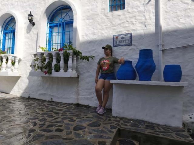 ZoeTobon69, with ass and tits, stands against a white wall, wearing a green cap and gray t-shirt, with blue pots and a signboard in a sunny, cobblestone alley.
