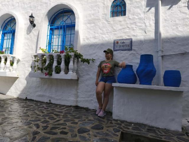 ZoeTobon69 with ass leaning on blue vases, wearing green tee, blue shorts, and cap, standing on cobblestone, white-washed wall with arched windows and flowers.