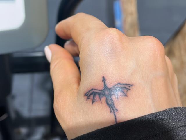 SteffanyTaylor's hand with a black bat tattoo on the back, wings spread, in a photo. Her fingers are slightly curled, showing white nails. She's gripping a car steering wheel.