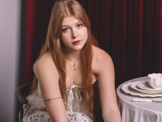 StellaLane with long red hair, fair skin, and small tits, wearing a white lace dress, leans forward on a chair, looking at the camera, with a table beside her.