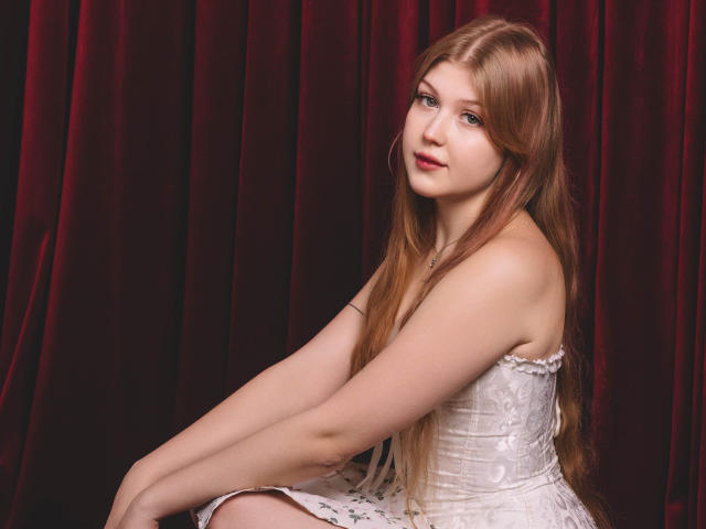 StellaLane, with long red hair, sits wearing a white, strapless, floral dress, showing her bare shoulders and arms, looking slightly to the side with a gentle smile.