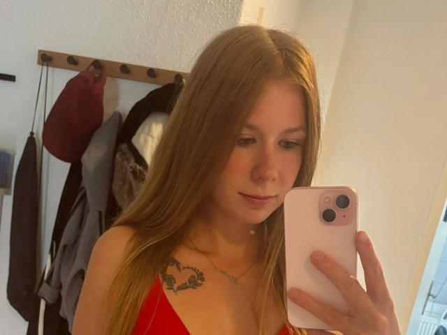 Soleea, attraktiv mit langen braunen Haaren, rotem Kleid, Tattoo auf der linken Brust, hält ein rosa iPhone, macht ein Selfie, leichtes Lächeln, weiches Licht, weiße Wände, rote und graue Hüte im Hintergrund..