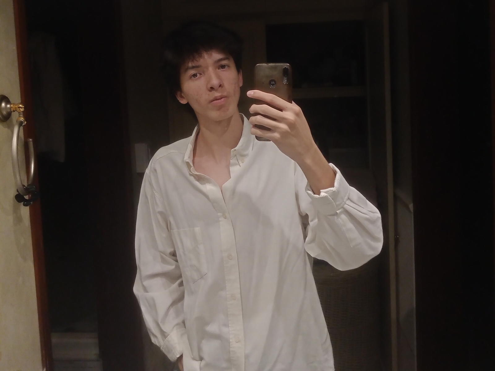 A selfie of GeniusMe shows a young man with short, dark brown hair and a neutral expression. He's wearing a loose, white, button-up shirt with his hands in his pockets. He's holding a gold-colored smartphone in his right hand, taking the photo in a dimly lit room. The background is dark, with a door on the left and shelves on the right. The image is slightly blurry, emphasizing the casual, everyday moment.