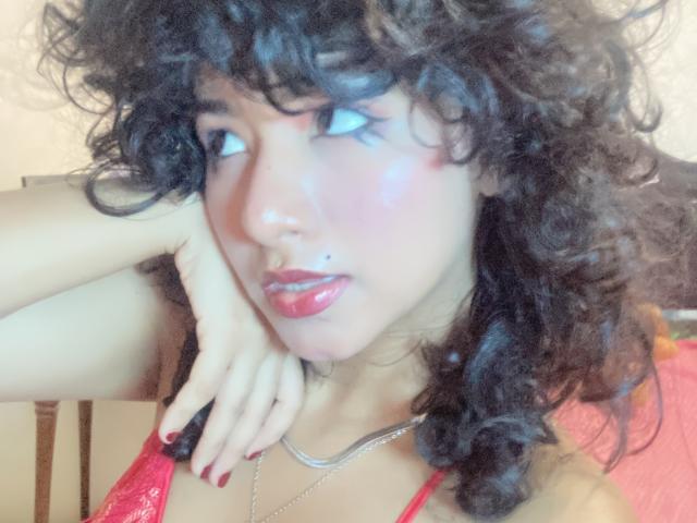 MargarethTorres has curly black hair, red lipstick, and a small mole on her upper lip. She's wearing a red lace top, with a silver necklace, and red nails.