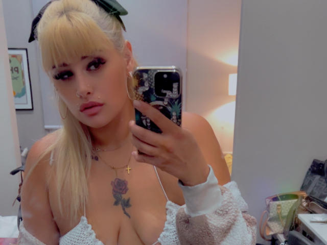 Tropicalbaddie69 with blonde hair in a ponytail, green cat ear bow, large boobs, rose tattoo, white off-shoulder top, holding phone in mirror selfie, soft pink lipstick.
