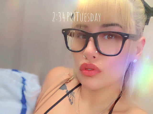 Tropicalbaddie69 with blonde hair, black glasses, nose ring, and pink lipstick, wearing a black top, showing a tattoo on her chest, and a subtle sheen on her face.