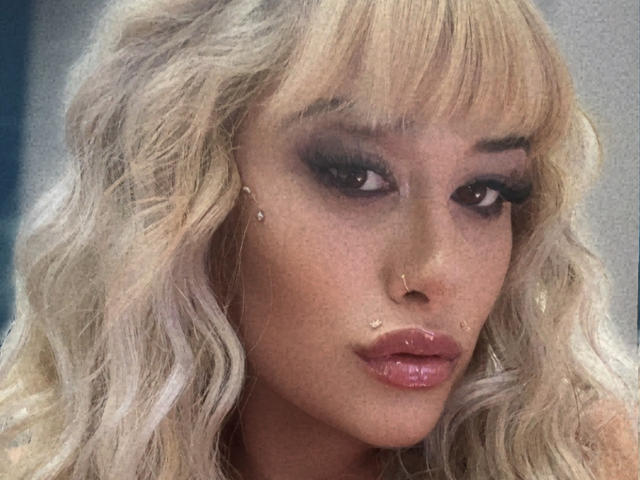 Tropicalbaddie69 has wavy blonde hair, dark smoky eye makeup, glossy pink lips, and small diamond studs on her cheeks. Her nose is pierced. She looks seductive.