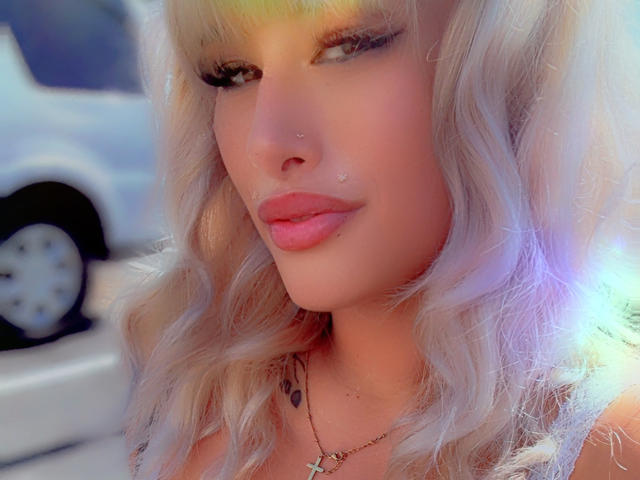 Tropicalbaddie69 has pale skin, pink lips, and wavy platinum blonde hair, with a nose piercing and subtle makeup, wearing a cross necklace, and a tattoo below the collarbone.