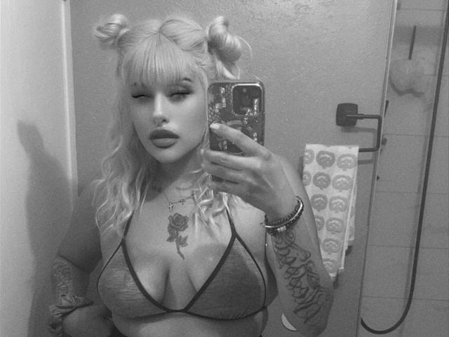 Black-and-white selfie of Tropicalbaddie69 with blonde pigtails, wearing a mesh bralette, showing tattoos on arm and chest, holding phone, visible rose tattoo on chest, large tits.