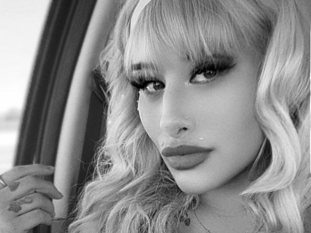Tropicalbaddie69 has blonde wavy hair, heavy eyelashes, and full lips with small beauty marks above her mouth. She's looking to the side with a slight pout, in black-and-white.