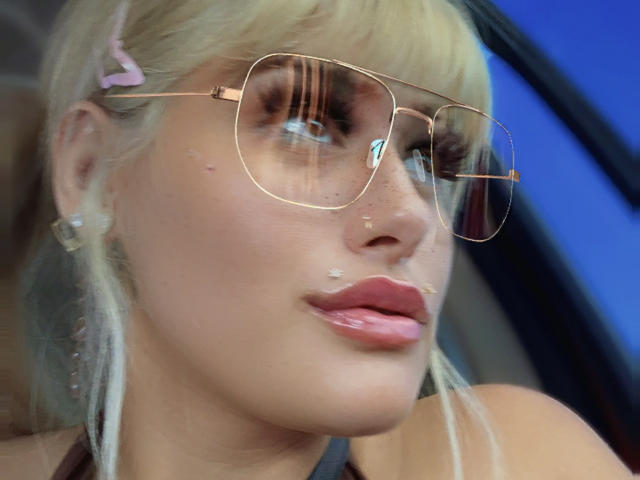 Tropicalbaddie69 with blonde hair, pink hairclip, gold aviator sunglasses, freckles, glossy pink lips, and a small beauty mark on her upper lip, looking to the side.