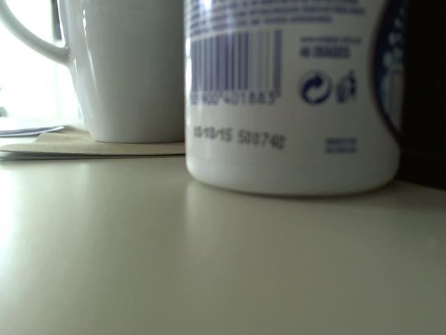 White ceramic mug with blue barcode and text, next to another white ceramic mug. Foreground shows a blurry white table. FlipFlops-hot's ass is not visible.