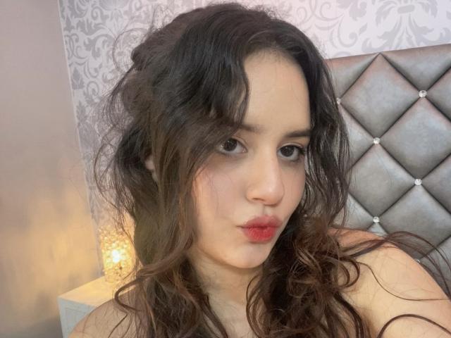 KylieLumi tiene el pelo castaño ondulado, ojos oscuros y lápiz labial rojo. Está haciendo un puchero ligeramente, con una mirada sexy y seductora, y sus pechos son parcialmente visibles..