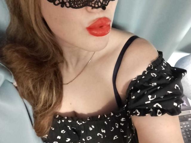 SofiaBells with red lips, black lace mask, wavy brown hair, black off-shoulder dress with white hearts, black strap, and necklace, showing her tits and upper body.