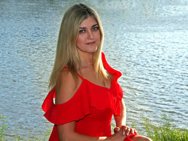 HelenUnique with blonde hair, red off-shoulder dress, and pearl necklace, sitting by a water body, smiling, showing her tits slightly, hands on her lap, greenery in front.