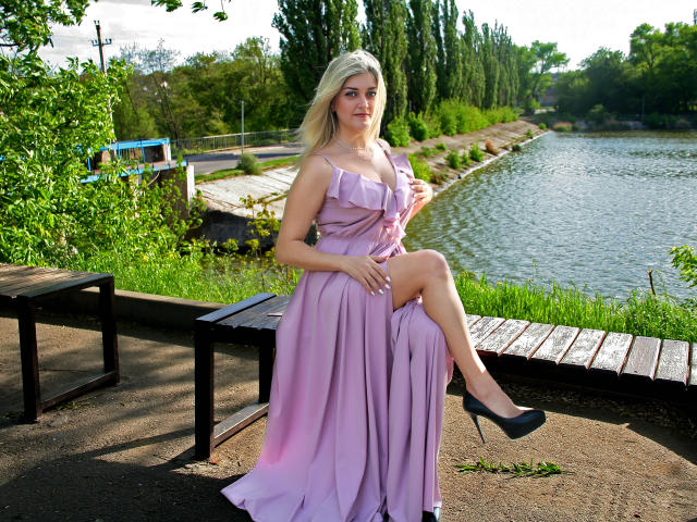 HelenUnique with pale purple dress, sitting on wooden bench by lake, legs crossed, black high heels, blonde hair, soft smile, green trees, water in background, natural light.
