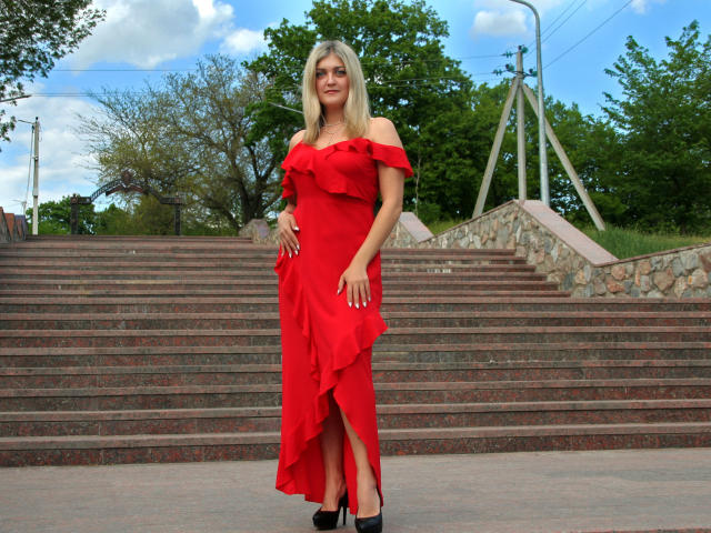 HelenUnique stands in a red off-shoulder dress with ruffled sleeves, a high slit, and black heels, smiling at the camera. Her blonde hair is straight and she's holding her ass.