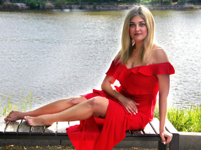 HelenUnique with blonde hair, off-shoulder red dress, sitting on wooden plank by water, legs crossed, bare feet, looking at camera, slight smile, hand on knee, daytime.
