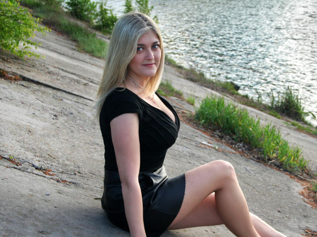 HelenUnique, blonde with medium-length hair, wearing a black dress and leather skirt, sitting on a concrete slope, showing her ass and small tits, with a silver necklace.