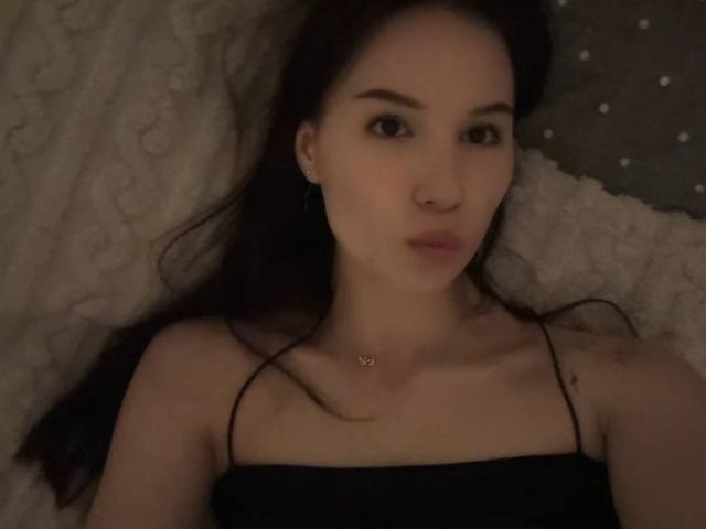 Amabel with dark hair, brown eyes, and a nose ring, lying down, wearing a black strap top, small tits, and a heart necklace, in dim lighting.