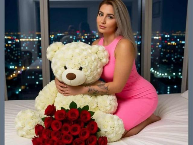 ChaudeEvely with medium tits, wearing a tight pink dress, kneeling on a bed, hugging a giant white teddy bear with red roses in front of her.