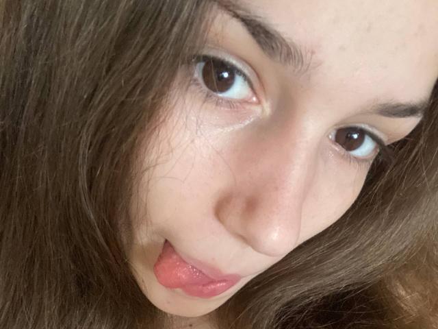 KaiaLein's face, brown eyes, and slightly parted pink lips with tongue out, brown wavy hair, fair skin, and thin eyebrows, close-up shot, looking up with a playful expression.