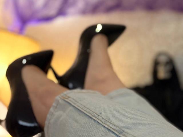 KaiaLein's legs in blue jeans with black high heels, feet pointed upward. Her shiny black shoes reflect light. Her legs are slightly tanned, relaxed.