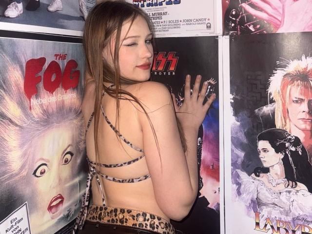 Bubochka with long brown hair, leopard print top, and backless dress, standing in front of horror movie posters, smiling with red lipstick, showing her back and small ass.