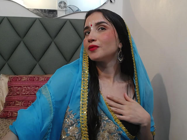 JalimaMona, avec ses longs cheveux noirs, sa robe brodée de bleu et d'or, son rouge à lèvres rouge, et ses boucles d'oreilles en argent, touchant sa poitrine, portant un bindi rouge, regardant vers le haut avec un léger sourire..