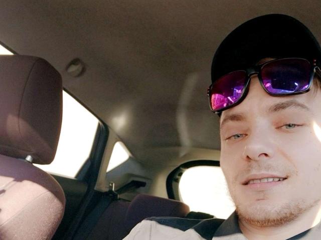 TedHott69 wearing a black cap, purple sunglasses, and a striped shirt, smiling with a slight beard, inside a car with grey headrests in the background.