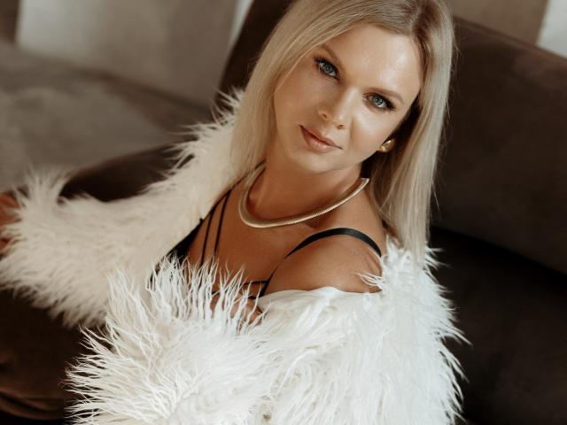 AgathaKlouin with blonde hair, blue eyes, and a cute face, wearing a black strap top, white fluffy jacket, and gold necklace, sitting on a brown couch.