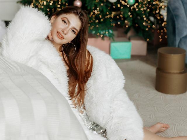 AmandaGasertoe sits on a beige carpet, wearing a fluffy white coat with large silver sequins. She has long brown hair, fair skin, and is smiling with her right arm behind her head. She has large hoop earrings. A Christmas tree with blurred golden lights and ornaments is in the background, along with a pink gift box and a brown cylindrical present. The foreground has a white, striped blanket. The image has a warm, festive atmosphere.